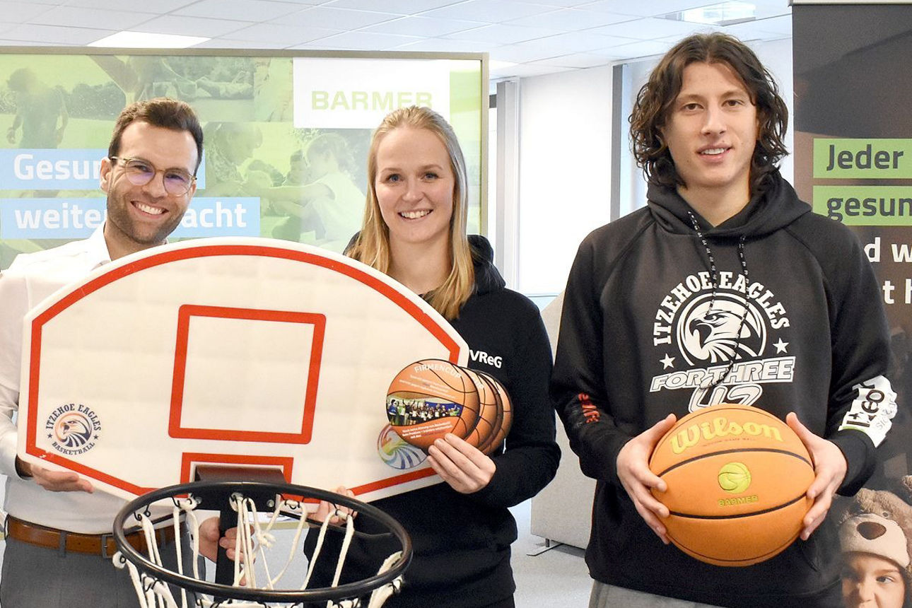 Vier Personen stehen lächelnd vor der Kamera. Die zwei mittleren Personen halten einen Basketballkorb..