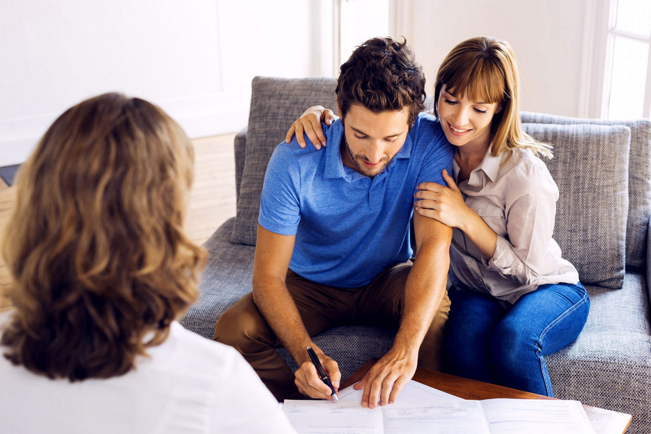 Junges Pärchen sitzt auf einem Zweisitzer-Sofa. Er unterschreibt Unterlagen. Sie umarmt ihn lächelnd.