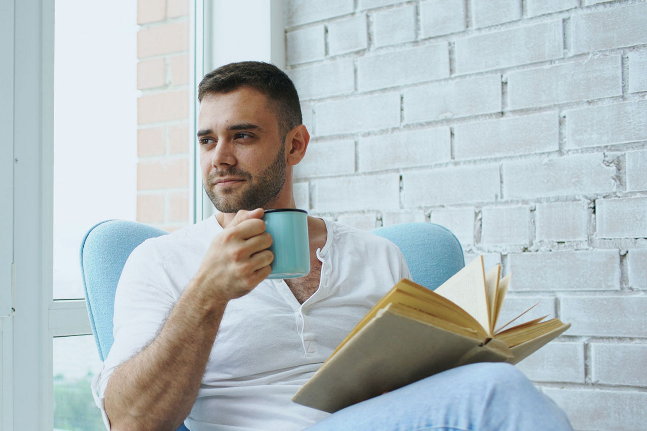 Ein Mann am Fenster auf seinem Sofa, der ein Buch und eine Tasse hebt.
