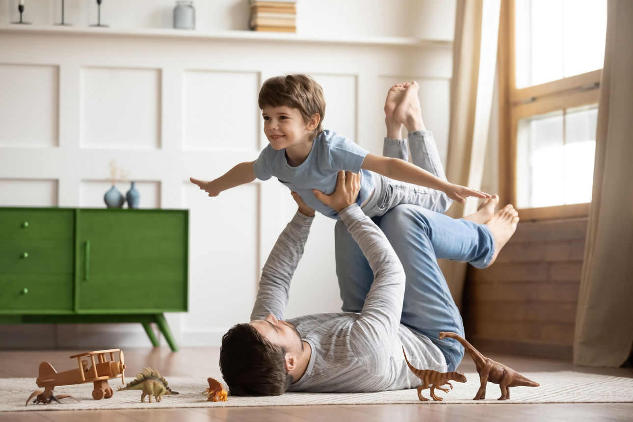 Ein Mann liegt auf dem Fußboden und hebt sein Kind wie ein Flugzeug in die Luft