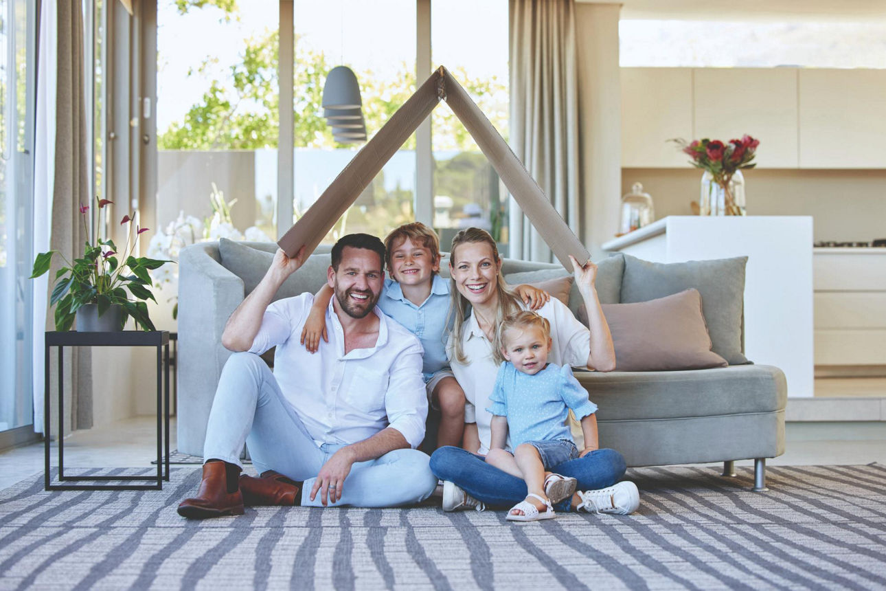 Eine Familie sitzt in ihrem Wohnzimmer und hält ein symbolisches Dach über ihren Kopf