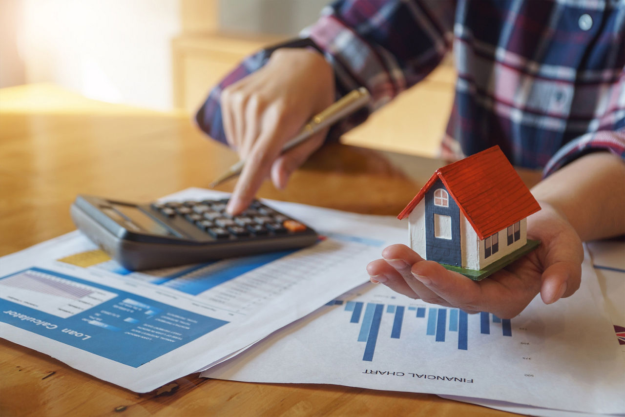 Eine Frau hält ein Modellhaus in der linken Hand. Mit der rechten Hand tippt sie auf einem Taschenrechner, der auf ein paar Blättern mit Diagrammen liegt.