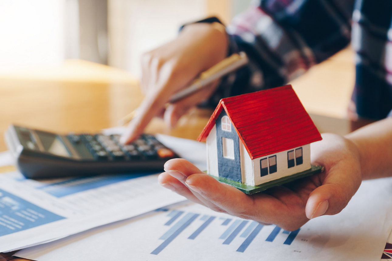 Eine Person, die an einem Taschenrechner rechnet und parrallel ein Miniaturhaus in der anderen Hand hält