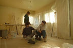 Eine Frau sitzt auf einem Hocker, rechts neben ihr sitzt ein Mann auf dem Boden. Im Hintergrund läuft eine weiter Frau. Sie befinden sich in einem Zimmer, das für Malerarbeiten vorbereitet wird. 
