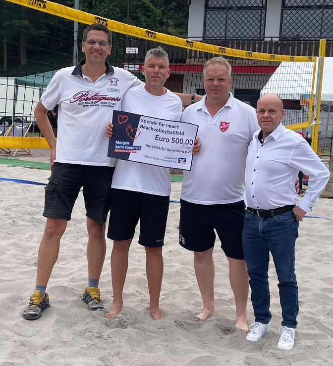 Vier Erwachsene stehen auf einem Sandplatz und halten einen Spendenscheck in der Hand.