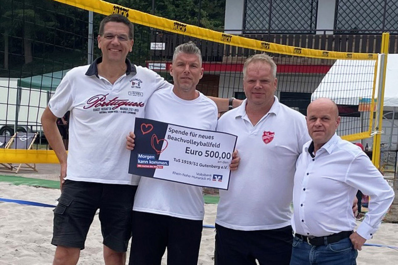 Vier Erwachsene stehen auf einem Sandplatz und halten einen Spendenscheck in der Hand.