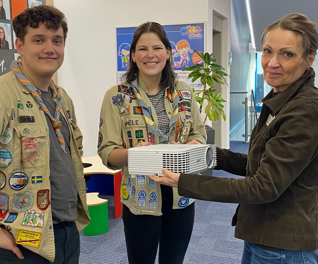 Drei Personen stehen in einem Innenraum zusammen und lächeln in die Kamera. Zwei von ihnen tragen Pfadfinderkleidung mit vielen Abzeichen. Gemeinsam halten sie einen weißen Beamer in der Hand. 