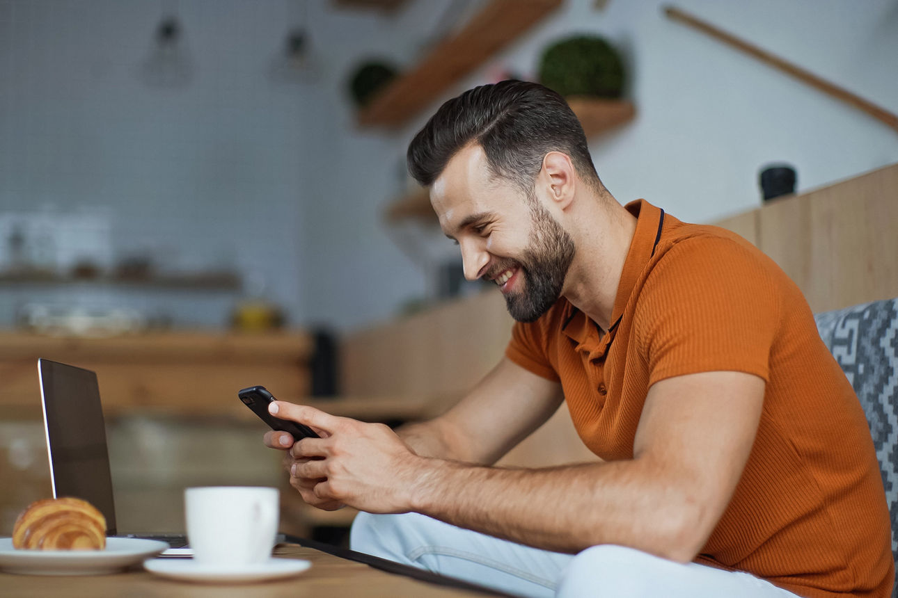 Ein Mann sitzt entspannt in einem Café, lächelt und schaut auf sein Smartphone. Vor ihm stehen ein Laptop, eine Tasse Kaffee 