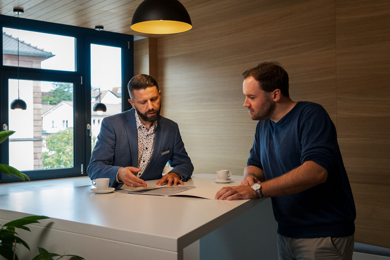 Ein Berater sitzt mit einem Kunden am Schreibtisch und sie trinken gemeinsam Espresso