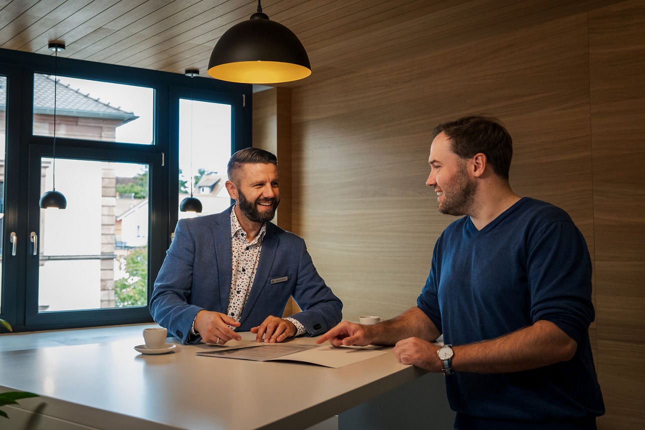 Ein Berater sitzt mit einem Kunden am Schreibtisch und sie trinken gemeinsam Espresso