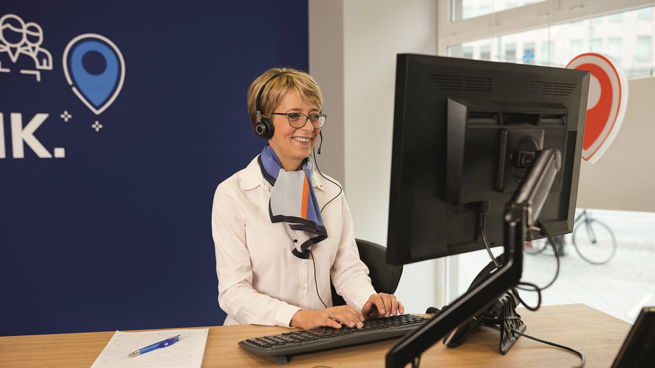 Frau mit kurzen blonden Haaren steht an einem Schreibtisch vor einem Monitor und trägt ein Headset