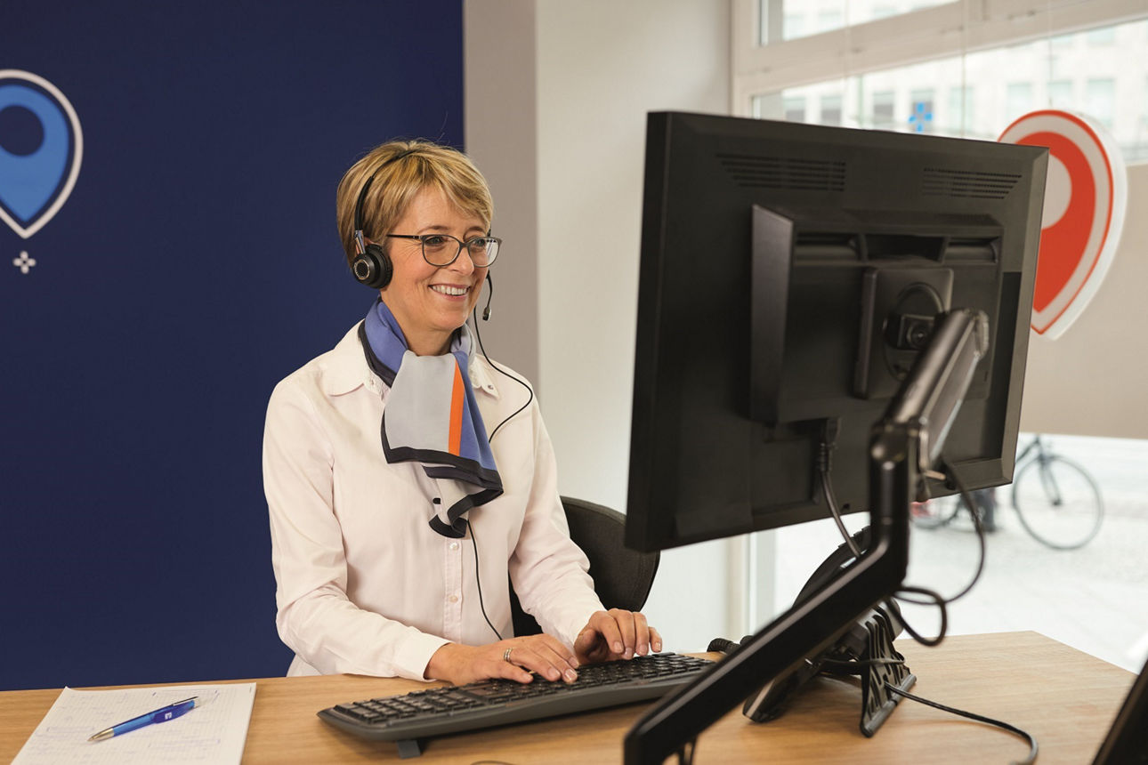 Beraterin mit weißer Bluse und Tuch sitzt mit Headset vor einem PC an einem Schreibtisch