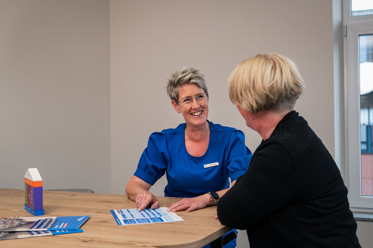 Eine Beraterin erklärt eine Kundin einen Flyer. Dabei sitzen die beiden an einem Tisch