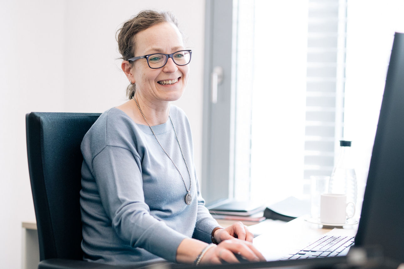 Lächelnde Frau mit Brille arbeitet an einem Computer in einem hellen Büro