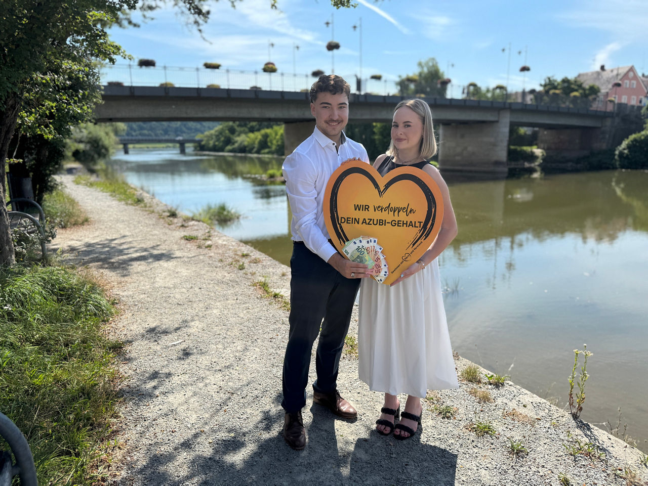 Ein junger Mann und eine junge Frau stehen an einem Fluss und halten Geld und ein Schild in der Hand
