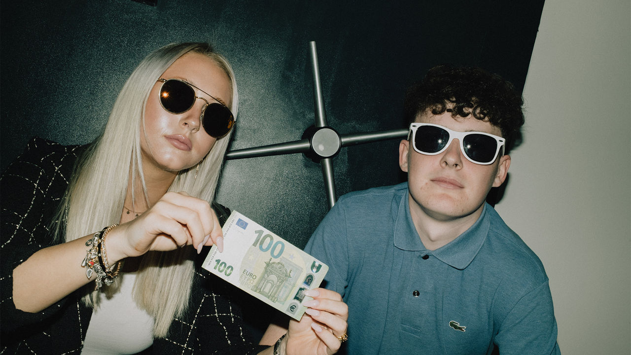 Zwei junge Menschen mit Sonnenbrillen vor einem Tresor, die junge Frau hält einen 100-Euro-Schein in der Hand.