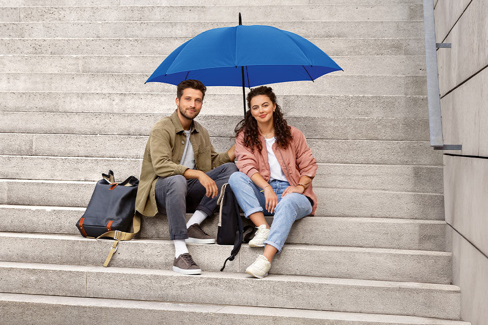 Junger Mann und junge Frau sitzen unter einem blauen Schirm auf einer Treppe und lächeln