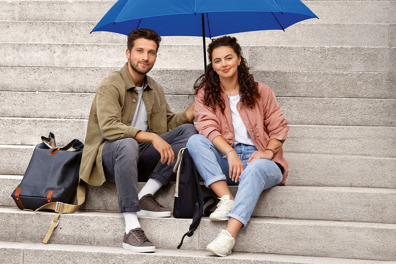Junger Mann und junge Frau sitzen unter einem blauen Schirm auf einer Treppe und lächeln