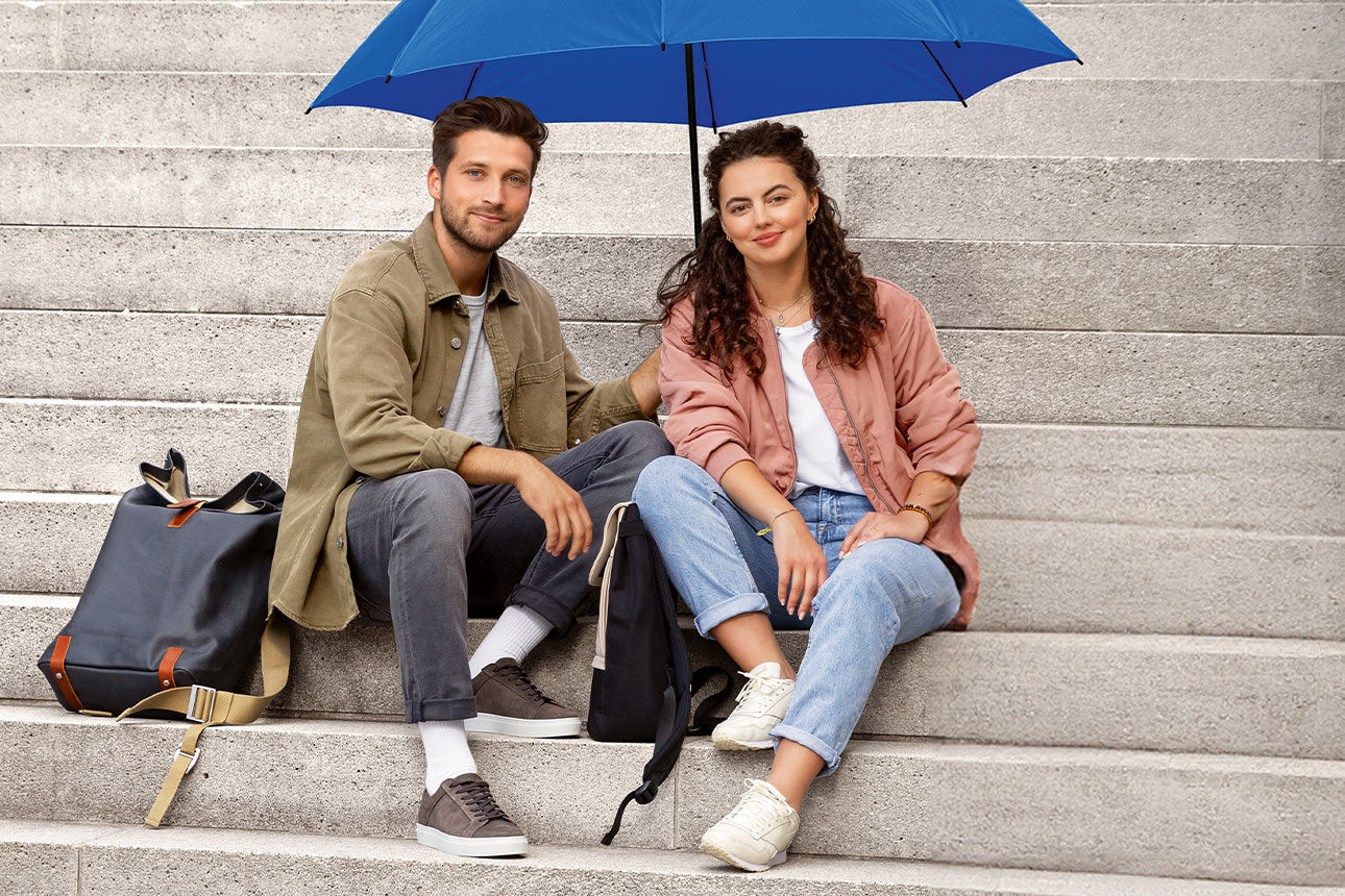 Junger Mann und junge Frau sitzen unter einem blauen Schirm auf einer Treppe und lächeln