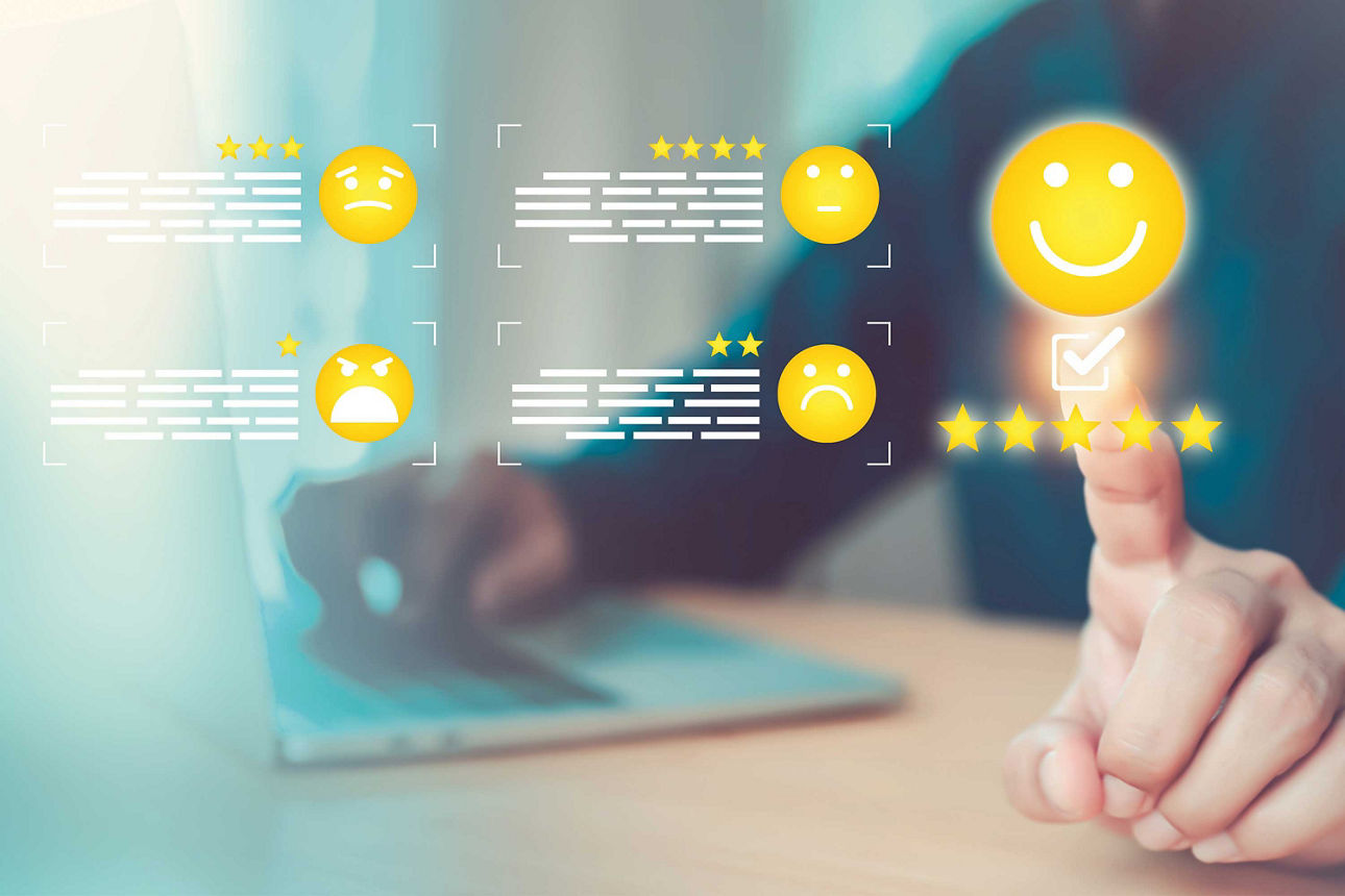 Hand tippt auf leuchtendes gelbes Smiley mit fünf Sternen, weitere gelbe Smileys mit Sternenbewertungen im Hintergrund, unscharfer Laptop auf Holztisch