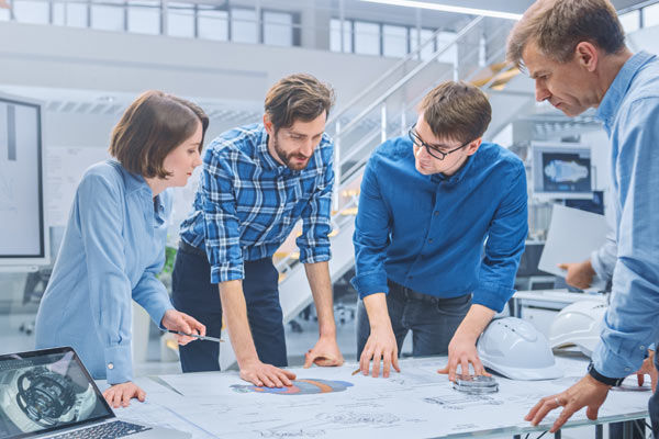 Vier Personen analysieren gemeinsam technische Zeichnungen an einem Tisch in einem modernen Büro