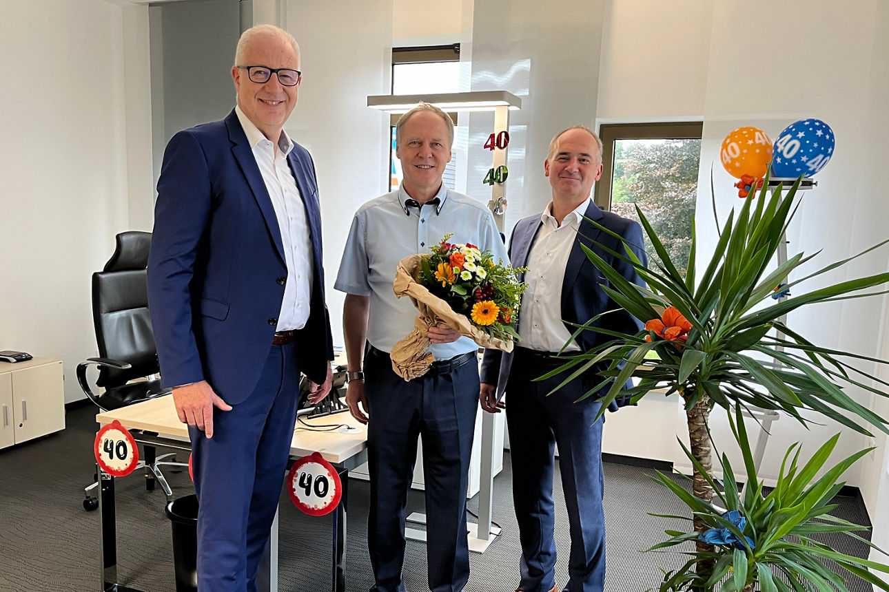 Drei Männer in der Mitte steht der Jublilar mit einem Blumenstrauß