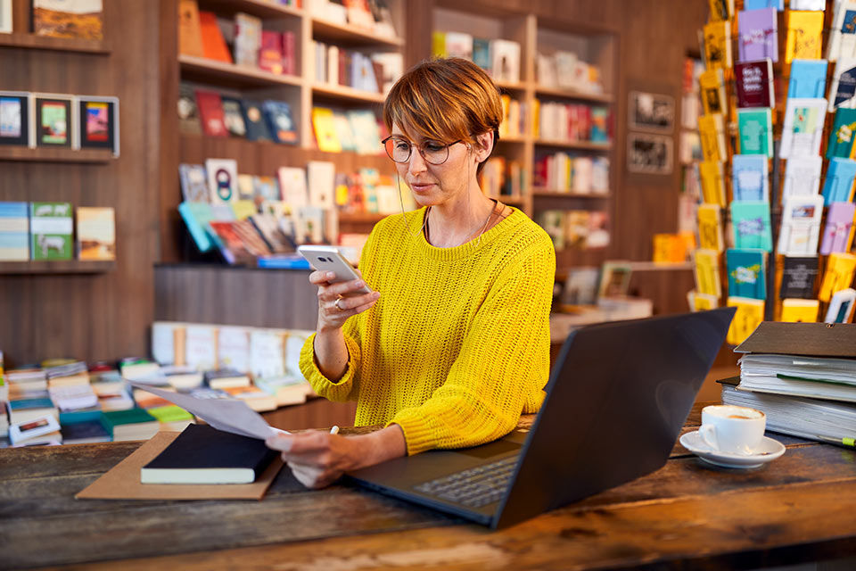 Eine Frau sitzt am Tisch vor einem Laptop mit Ihrem Handy in der Hand