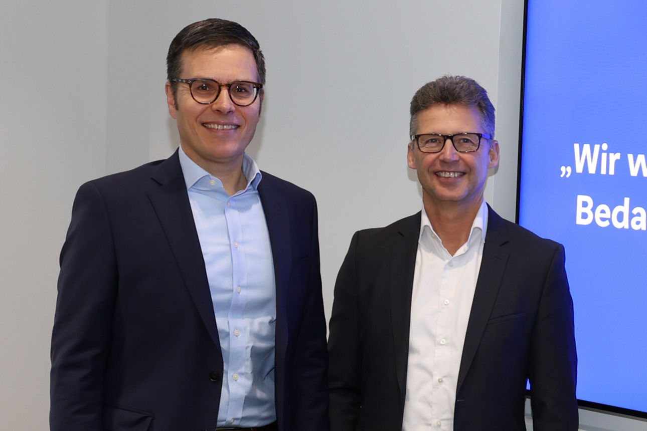 ie Vorstände Christoph Gommans und Stefan Rinsch stehen lächelnd vor einem Bildschirm mit Präsentationstext.