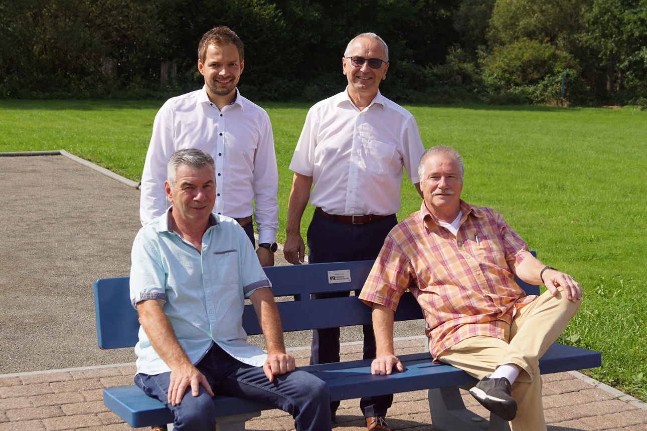 Vier Männer stehen und sitzen auf einer blauen Bank in einem Park