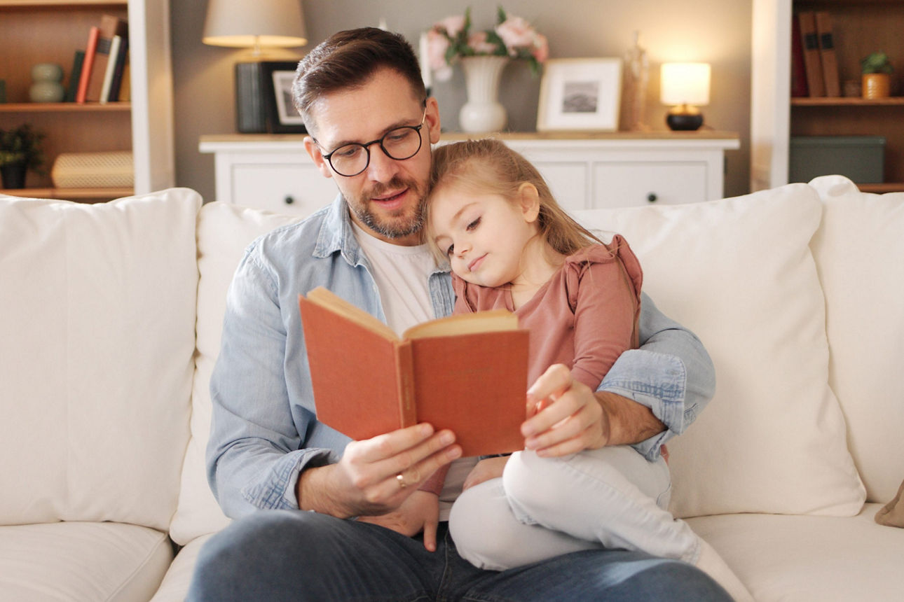 Vater liest seiner jungen Tochter ein Buch vor während sie auf dem Sofa entspannen