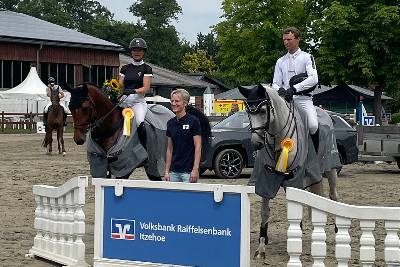 Eine Frau, die zwischen zwei Reitern auf einem Reitplatz im Freien steht.