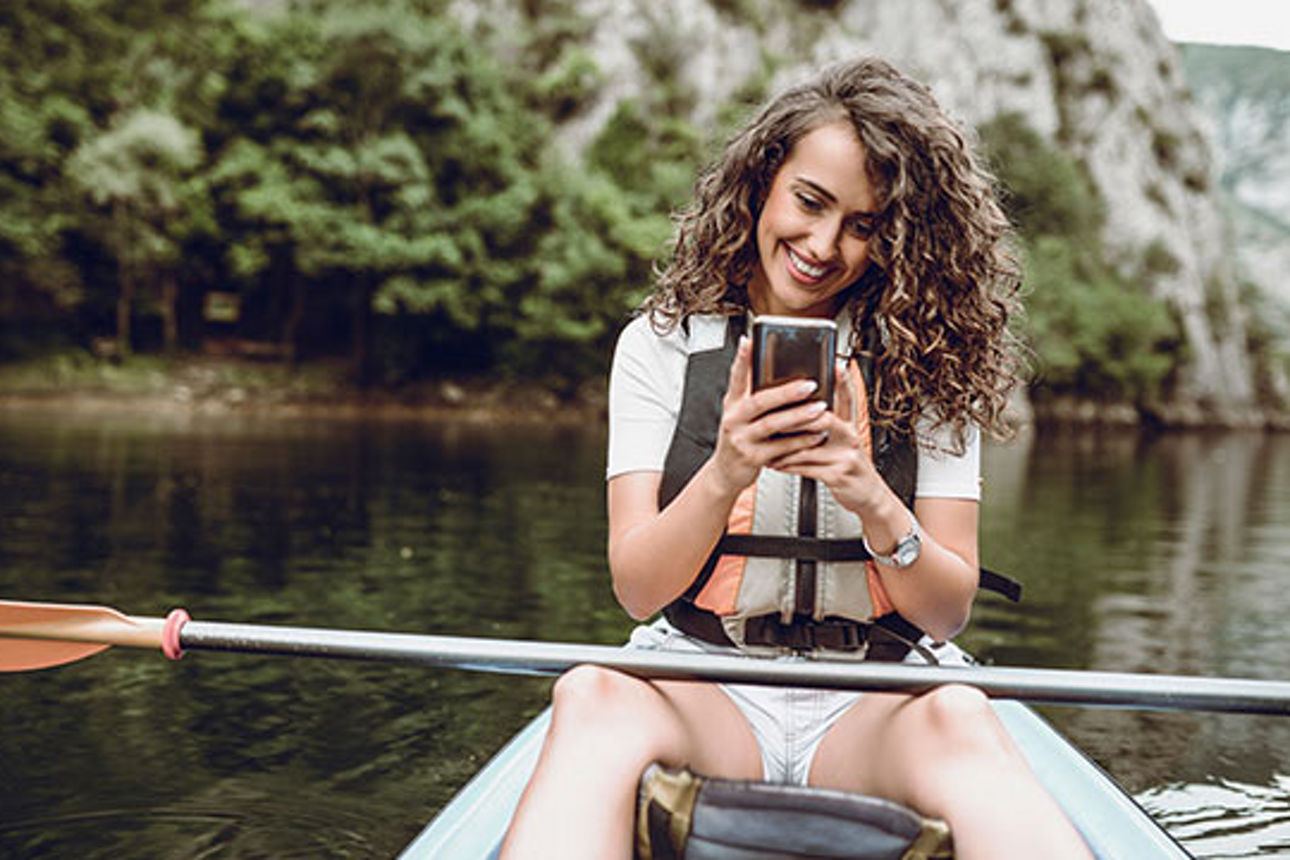 Junge Frau sitzt in einem Boot, mit Paddel auf den Beinen, und schaut lachend auf Ihr Handy