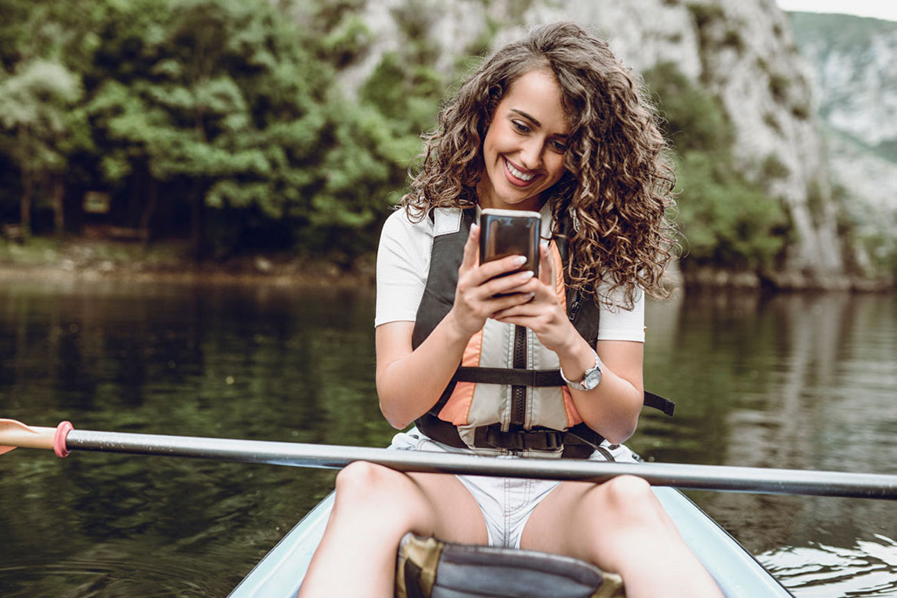 Junge Frau mit sitzt in einem Boot, das Paddel auf den Beinen, und schaut lachend auf ihr Smartphone