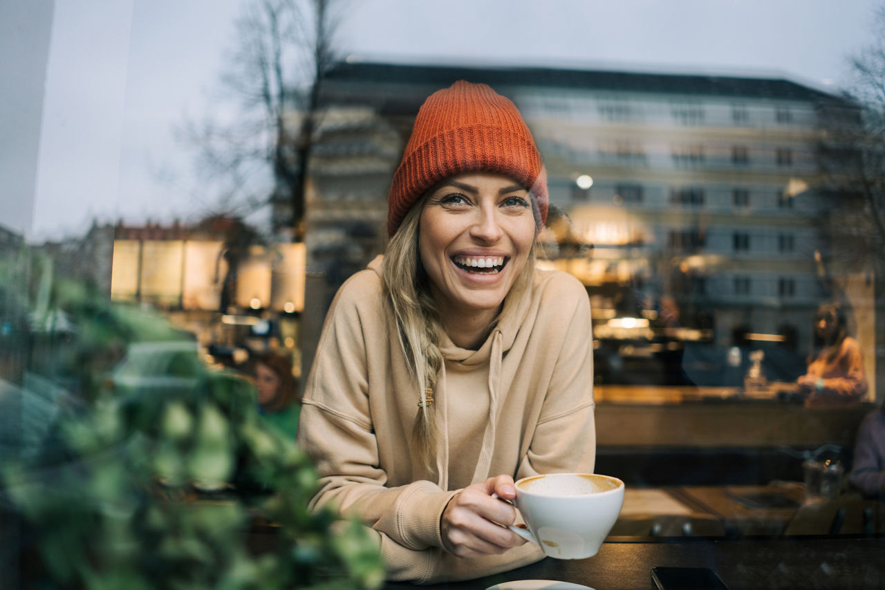 Foto von einer jungen, blonden Frau mit einer Kaffeetasse in der Hand, die lächelnd aus dem Fenster schaut