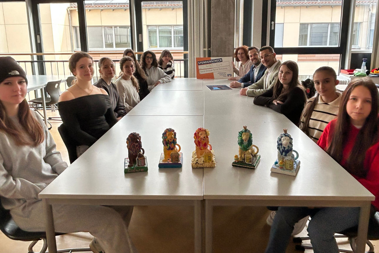 Gruppe Jugendlicher sitzt an einem langen Tisch mit mehreren bunten Figuren, im Hintergrund Erwachsene mit Spendenscheck.