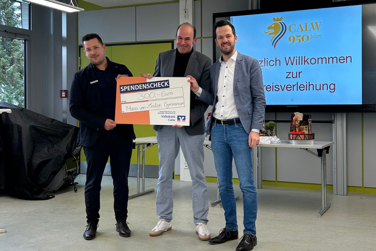 Drei Erwachsene präsentieren einen Spendenscheck vor Bildschirm mit Jubiläumslogo, auf dem Tisch stehen dekorative Figuren.