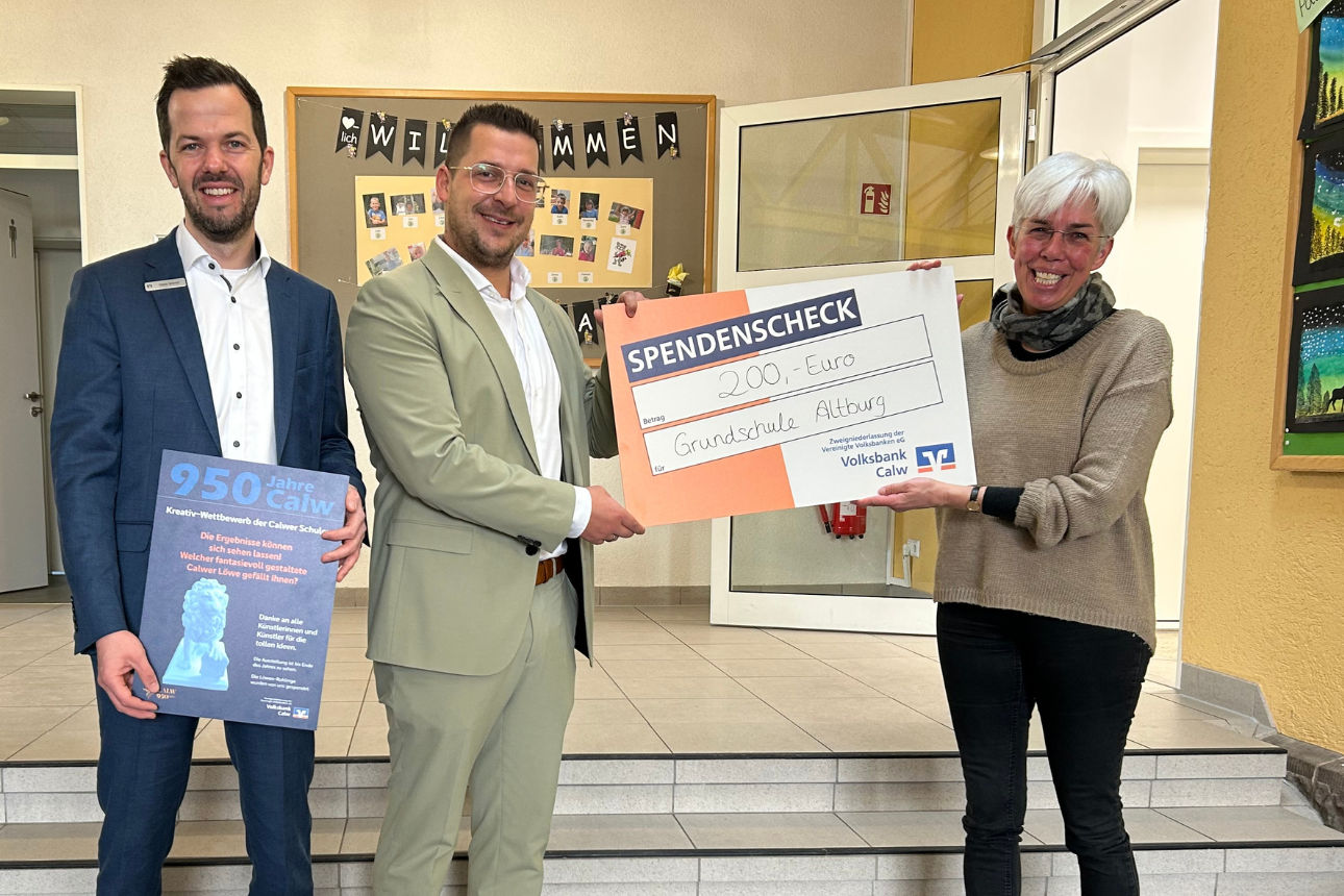 Drei Erwachsene überreichen in einem Schulflur einen Spendenscheck, daneben ein Plakat zum Jubiläum der Stadt Calw.