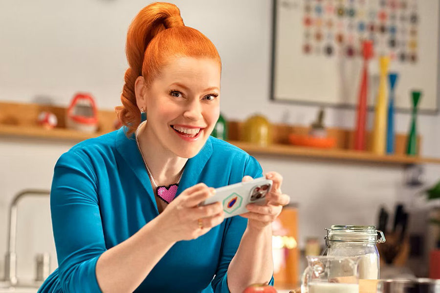 Enie van de Meiklokjes sitzt mit einem Handy in der Hand an einem Küchentisch und lächelt in die Kamera.