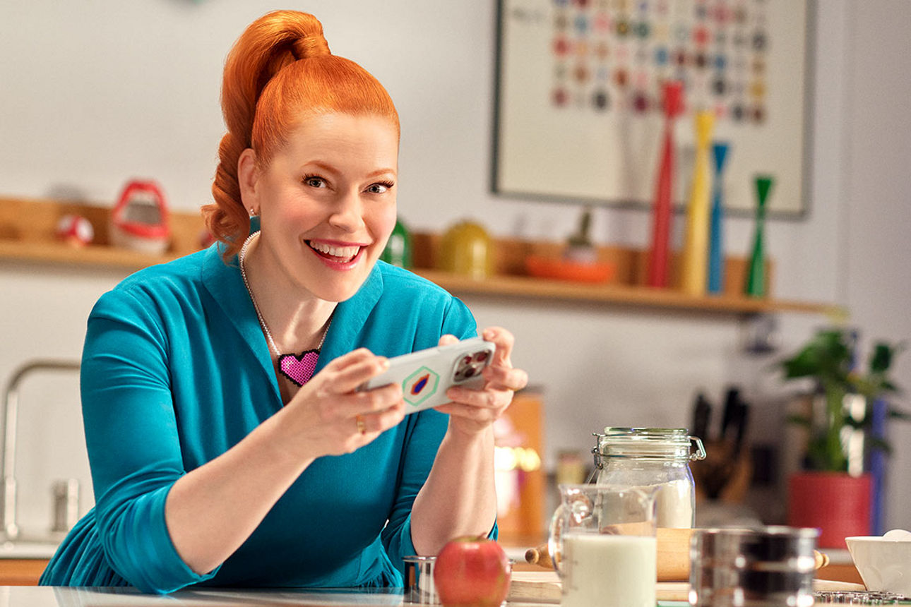 Enie van de Meiklokjes sitzt mit einem Handy in der Hand an einem Küchentisch und lächelt in die Kamera.