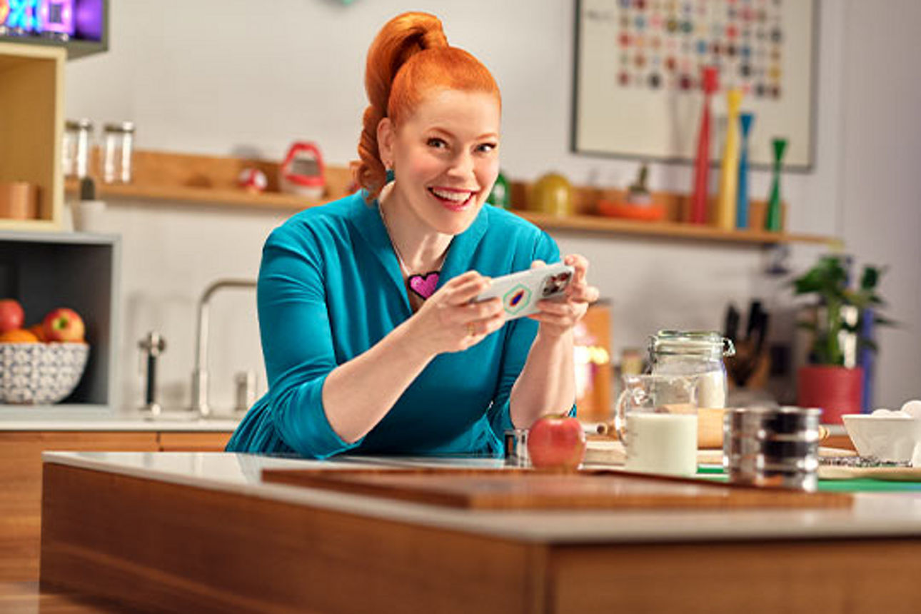 Eni van de Meiklokjes sitzt mit Handy in der Hand an einem Küchenmtisch und lächelt in die Kamera.