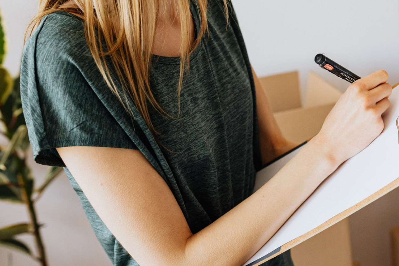 Frau hält Klemmbrett in  der Hand und macht Notizen. Im Hintergrund sind Umzugskartons zu sehen.