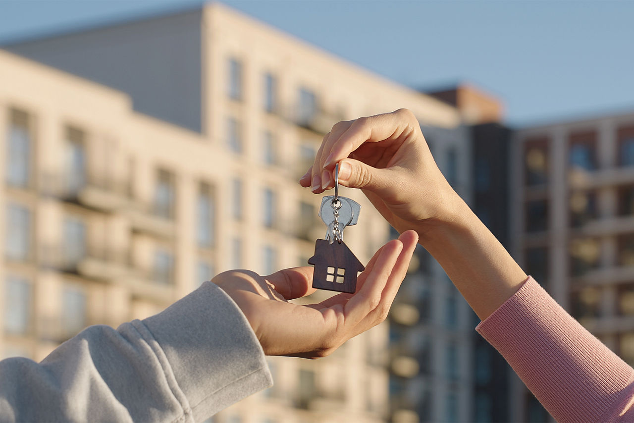 Nahaufnahme einer Frauen Hand die einen Schlüssel an an eine Männerhand übergibt. Im Hintergrund sieht man verschwommen ein Hochhaus