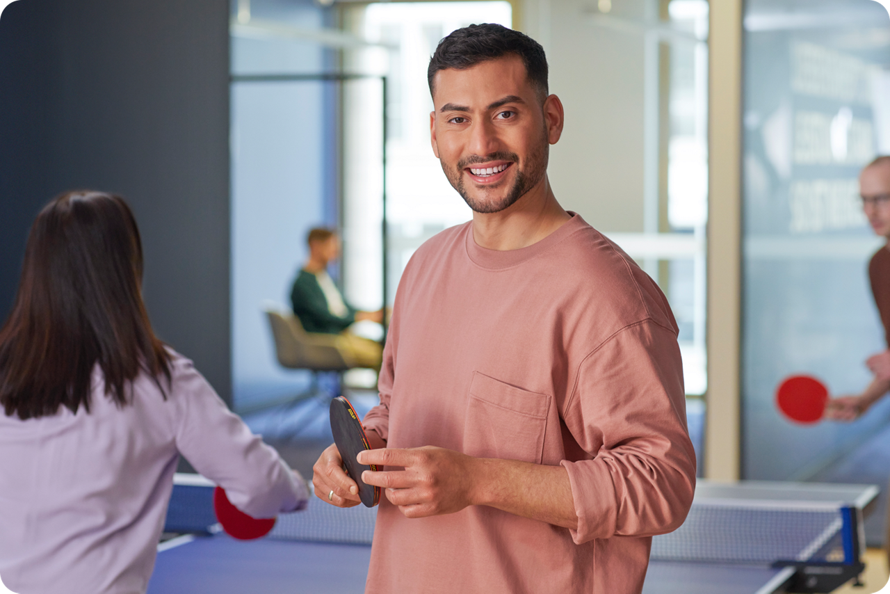 Ein junger Mann steht vor einer Tischtennisplatte mit einem Schläger in der Hand und lächelt in die Kammera, im Hintergrund w