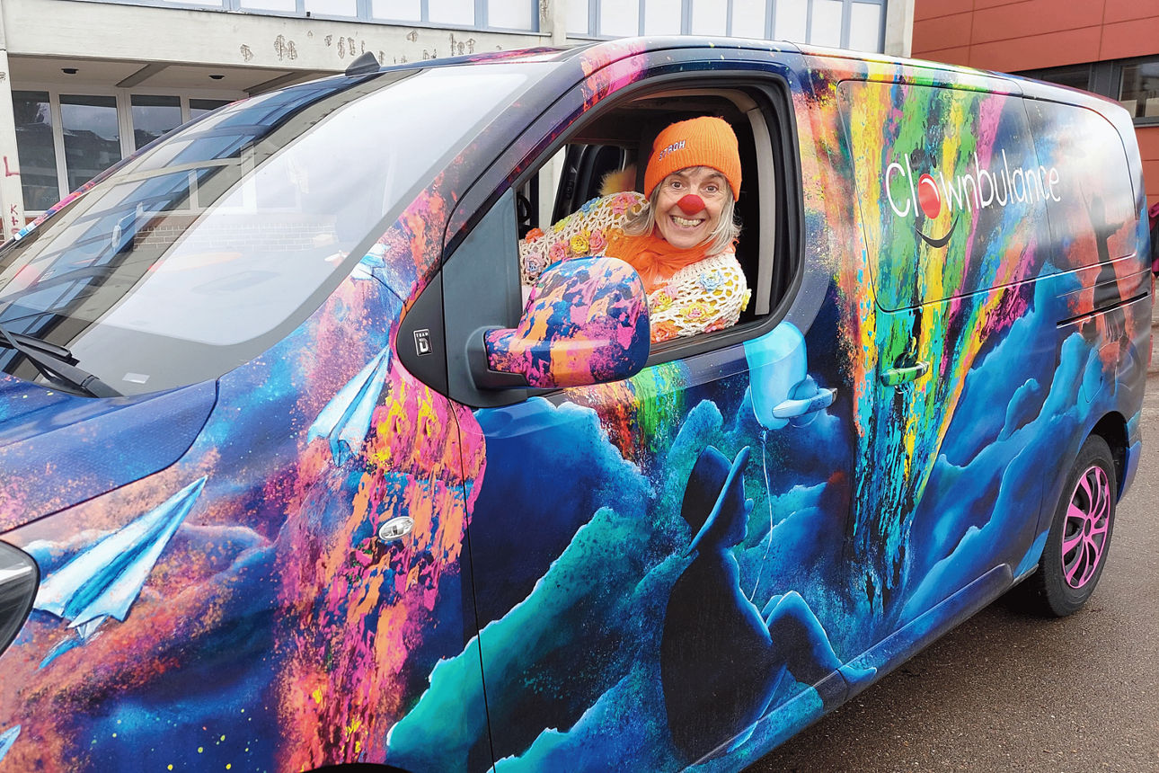 Eine Frau als Clown verkleidet im bunten Clownbulance-Fahrzeug. 