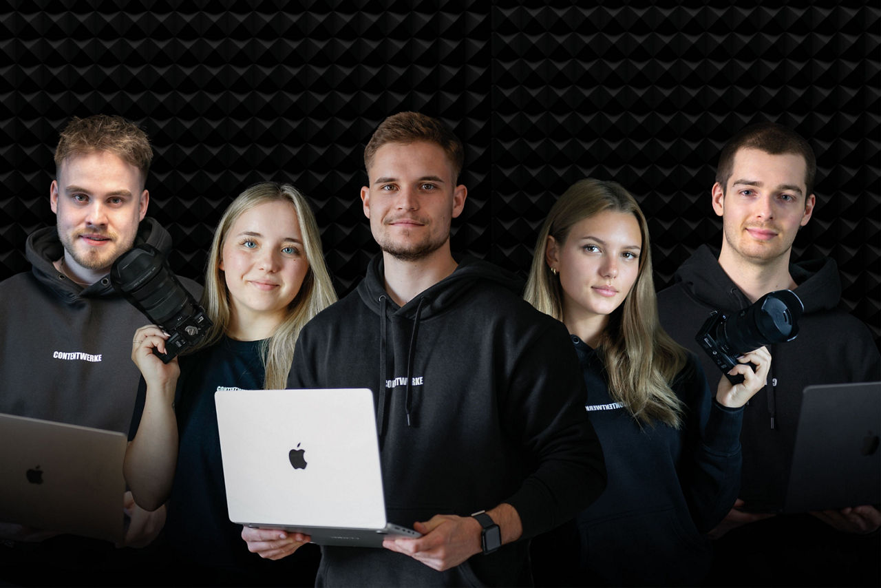 5 Personen stehen vor schwarzem Hintergrund, davon halten zwei eine Kamera in der Hand und drei einen Laptop.