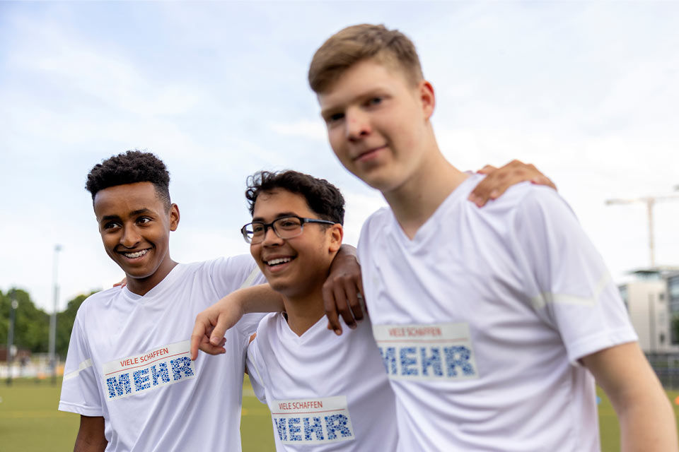 Auf einem Sportplatz stehe drei Jungen in Trikots nebeneinander