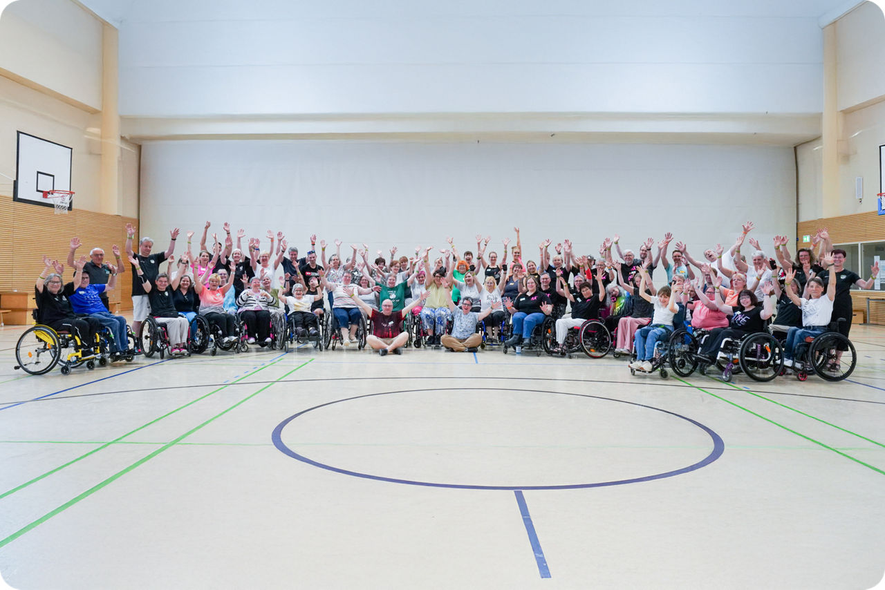 Große Gruppe von Menschen, viele im Rollstuhl, in einer Sporthalle mit erhobenen Armen und lächelnd in die Kamera.