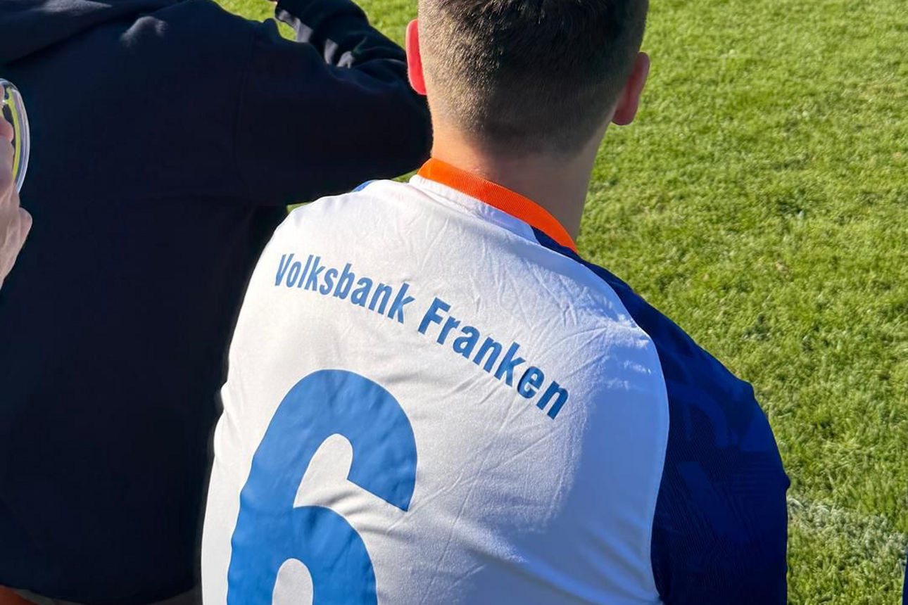 Blick von der Bank auf das Spielfeld; Rücken eines Spielers mit Aufdruck „Volksbank Franken“, Spiel läuft im Hintergrund.
