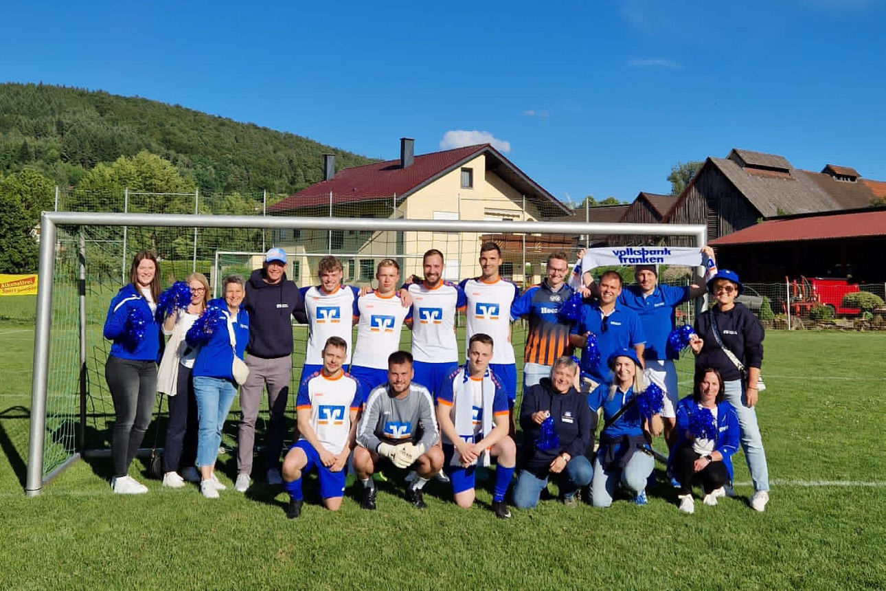 Team und Fans mit blauen Pompons posieren vor dem Tor auf dem Rasen, sonniger Himmel.