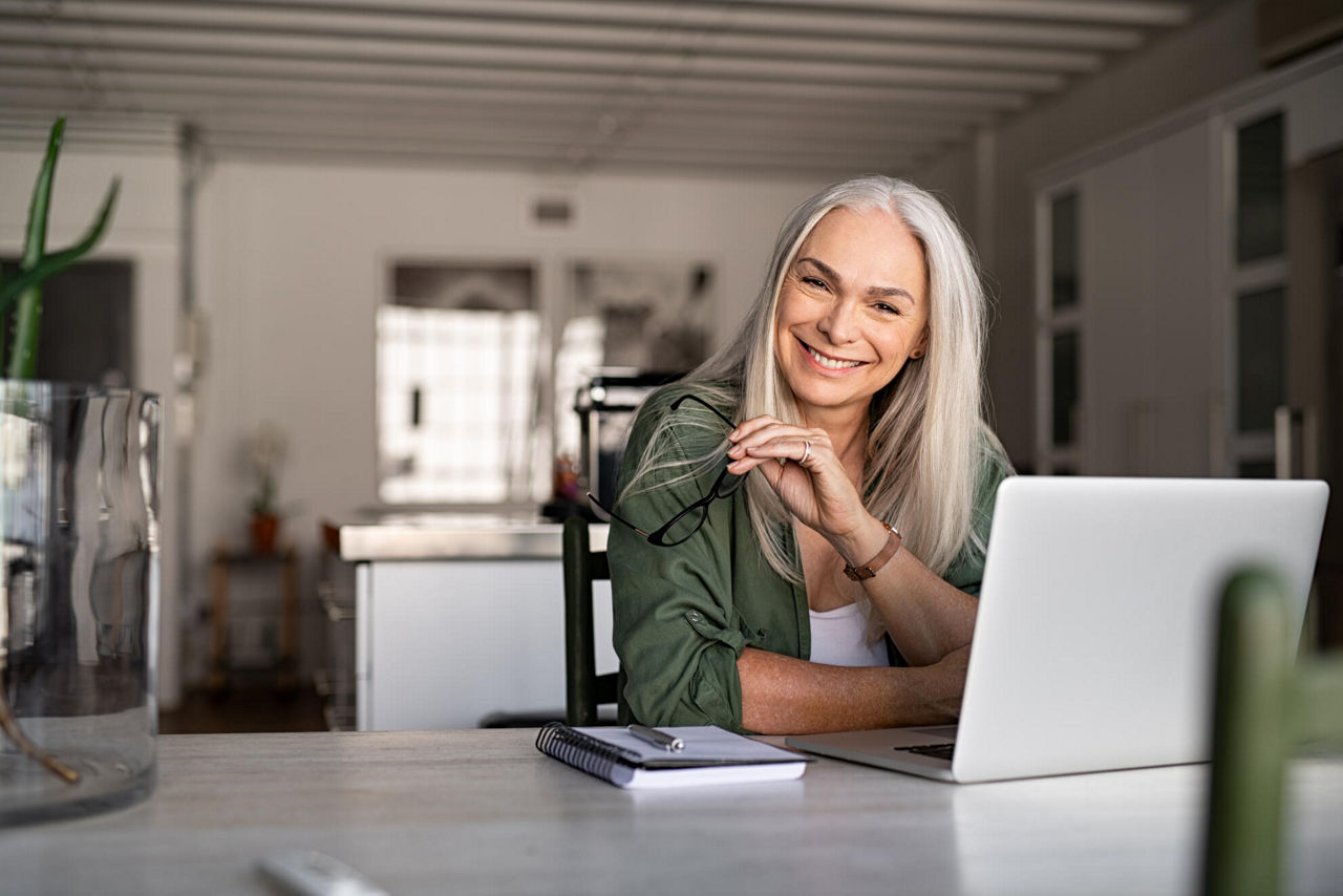 Eine ältere Dame sitzt zu Hause vor ihrem Laptop. Sie hält die Brille in der Hand und lächelt.